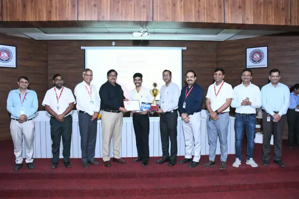 A row of about ten faculty members standing on stage with an ATF fellow, holding a trophy and a certificate between them