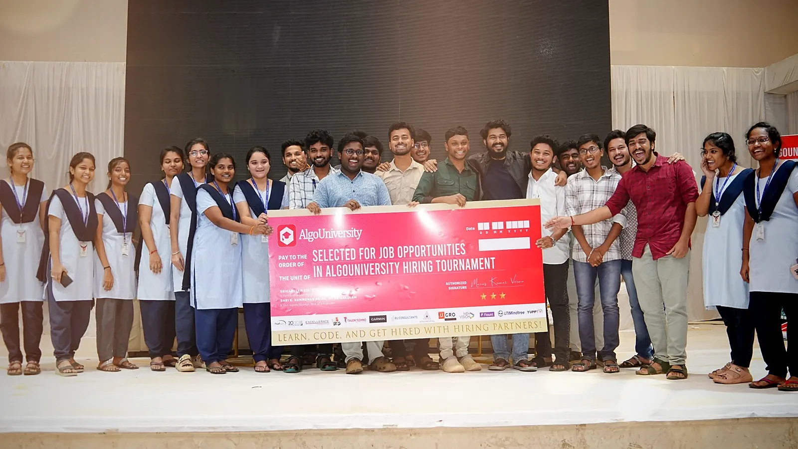 About twenty students on stage holding a large ceremonial cheque reading Selected for job opportunities in the AlgoUniversity hiring tournament