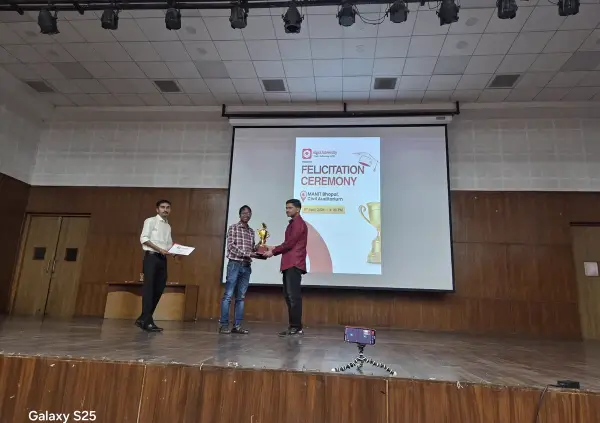 A fellow being handed a trophy on stage in front of a projected ATF felicitation ceremony slide at MANIT Bhopal