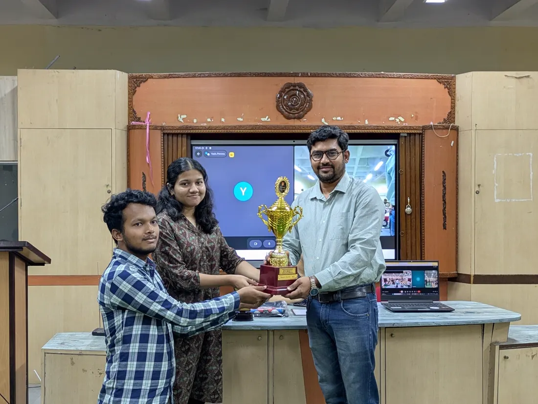 Two students receiving a trophy from a faculty member on a wooden stage